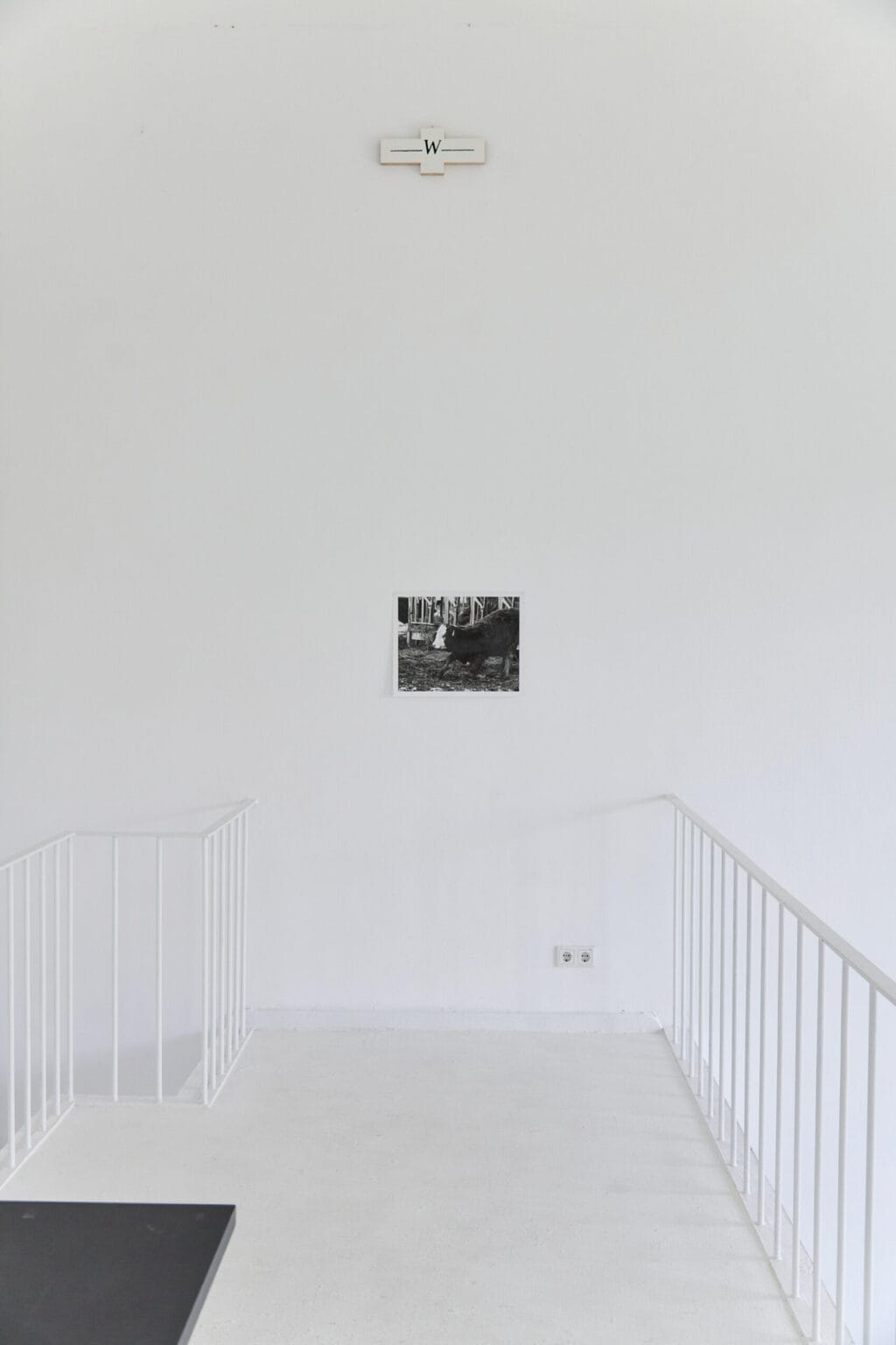 Foto: Eine schmale Empore mit Geländern, weiße Wände und weißer Boden. An der Wand hängen, auf unterschiedlicher Höhe mit viel Abstand untereinander, zwei Objekte.