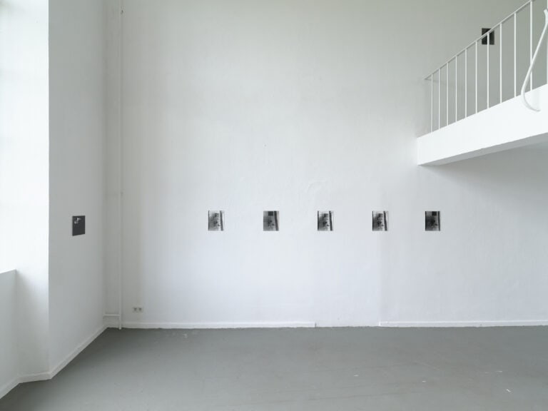 Foto: Aufnahme eines Ausstellungsraums mit weißen Wänden und grauem Boden. Auf der rechten Seite ist eine Empore zu erkennen. An der Wand sind sieben kleinformatige rechteckige Bilder angebracht.