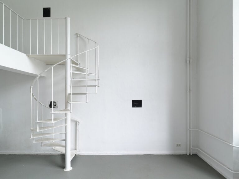 Foto: Aufnahme eines Ausstellungsraums mit weißen Wänden und grauem Boden. Auf der linken Seite ist eine Empore mit einer Wendeltreppe zu erkennen. An der Wand sind drei kleinformatige rechteckige Bilder angebracht.