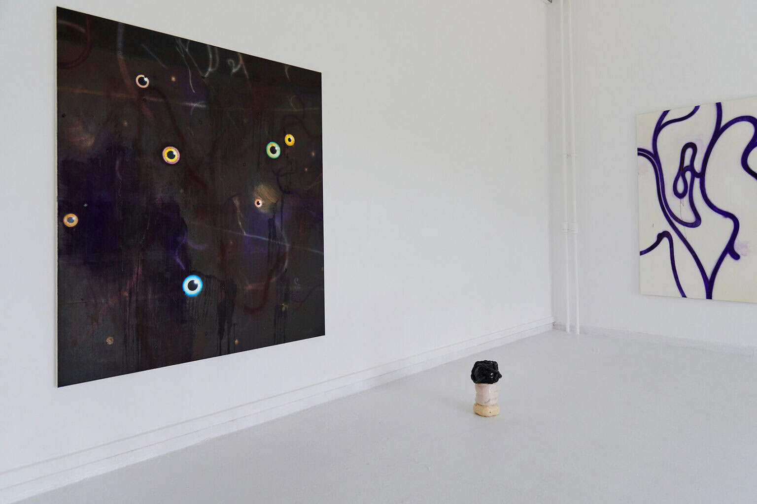 Foto: Die Ecke eines Raumes mit weißen Wänden und weißem Boden. An den Wänd hängen großformatige Leinwandarbeiten. Auf dem Boden steht ein Objekt.