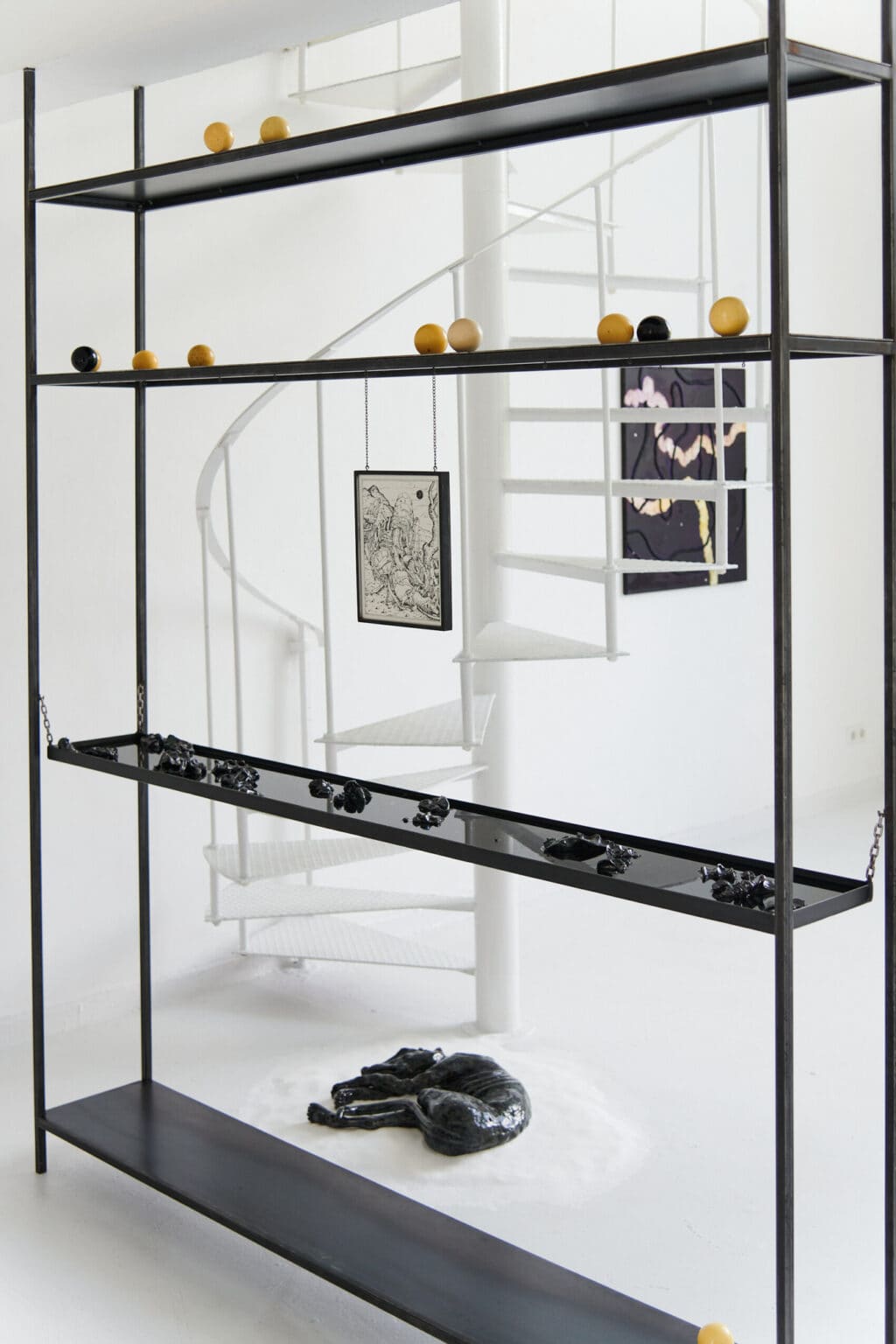 Foto: Ein schwarzes Regal mit vier Ebenen in einem Raum mit weißen Wänden, weißem Boden und weißer Wendeltreppe. In dem Regal befinden sich Objekte und Flüssigkeit. Von einer der Regalebenen hängt ein Objekt an Ketten herab. Auf dem Boden liegt ein glänzendes Objekt auf einem Haufen weißem Sand auf dem Boden.