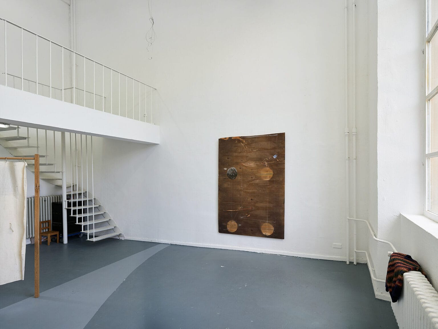 Foto: Ein Raum mit weißen Wänden, grauem Boden, einem Fenster und einer durch eine Treppe begehbaren zweiten Ebene mit Geländer.An der Wand hängt und im Raum steht ein Objekt. Auf den Boden ist in einem helleren Grauton eine From aufgemalt.