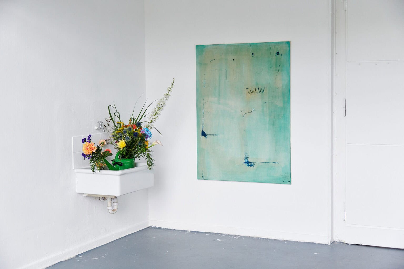 Foto: Ecke eines Raumes mit weißen Wänden. An der Wand hängt eine großformatige Leiwandarbeit. Daneben ein Waschbecken, in dem sich Blumen befinden.