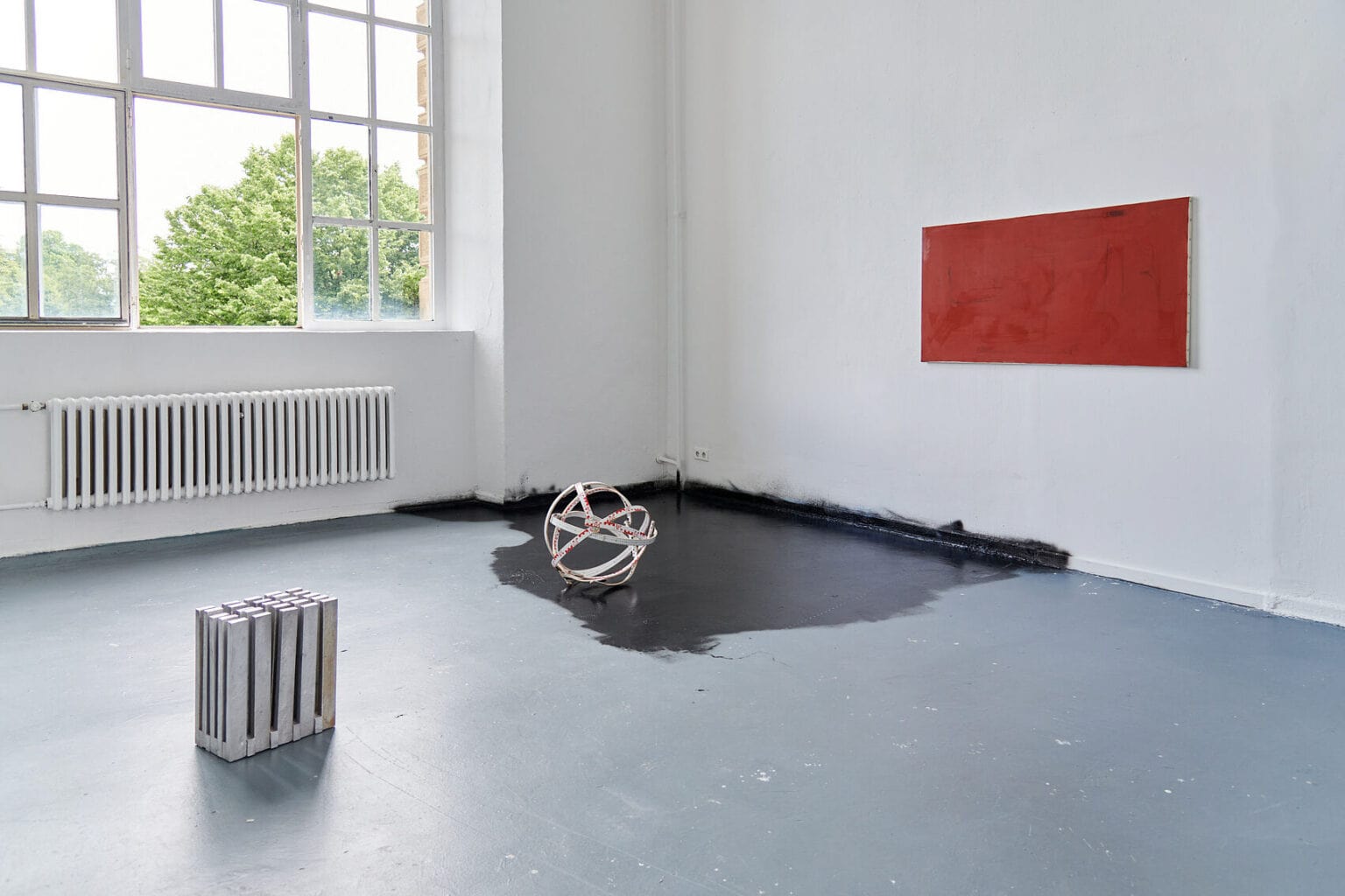 Foto: Ein Raum mit weißen Wänden, grauem Boden und einem Fenster. Eine Ecke des Raumes (der Boden, die Fußleisten und kleine Teile der Wand) sind mit schwarzer Farbe bemalt, die in unregelmäßigen Kanten ausfaded. Auf dem Boden liegen zwei Objekte. An der Wand hängt eine Leinwand im Querformat.