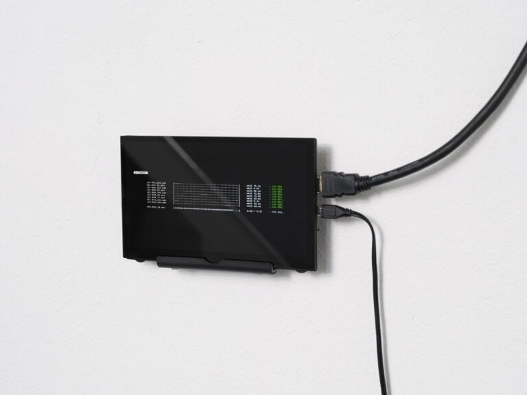 Foto: Detailaufnahme eines kleinen schwarzen Displays, welches an einer weißen Wand angebracht ist. Rechts im Gerät sind zwei Kabel eingesteckt.