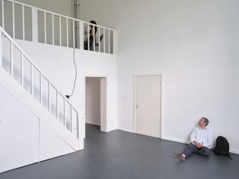 Foto: Aufnahme eines Ausstellungsraums mit weißen Wänden und grauem Boden. Auf der linken Seite ist eine Empore mit Treppenaufgang zu erkennen. An der Wand des Treppenaufgangs ist ein kleines schwarzes Gerät befestigt mit Kabelführung nach oben und unten. Auf dem Boden an der Rückwand sitzt eine Person. Auf der Empore sitzt eine Person auf einem Stuhl und spielt Saxophon.