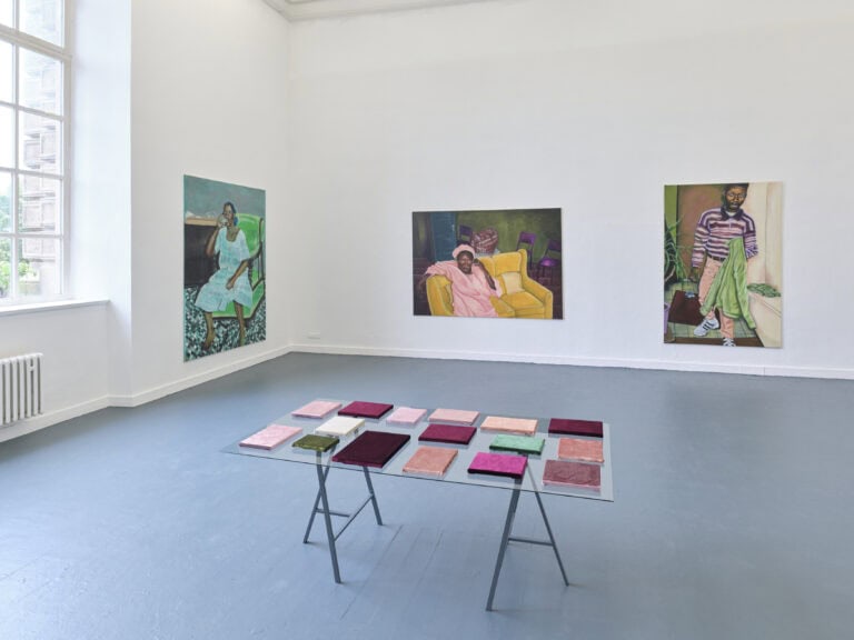 Foto: Aufnahme eines Ausstellungsraums mit weißen Wänden und grauem Boden und Fenster. An den Wänden sind drei großformatige Gemälde gehängt. In der Mitte des Raums steht ein Tisch auf dem bunte rechteckige flache Elemente zu sehen sind.
