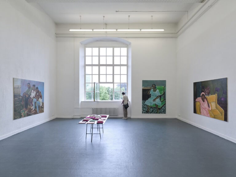 Foto: Aufnahme eines Ausstellungsraums mit weißen Wänden und grauem Boden und Rundbogenfenster. An den Wänden sind drei großformatige Gemälde gehängt. In der Mitte des Raums steht ein Tisch auf dem bunte rechteckige flache Elemente zu sehen sind. Am Fenster steht eine Person.