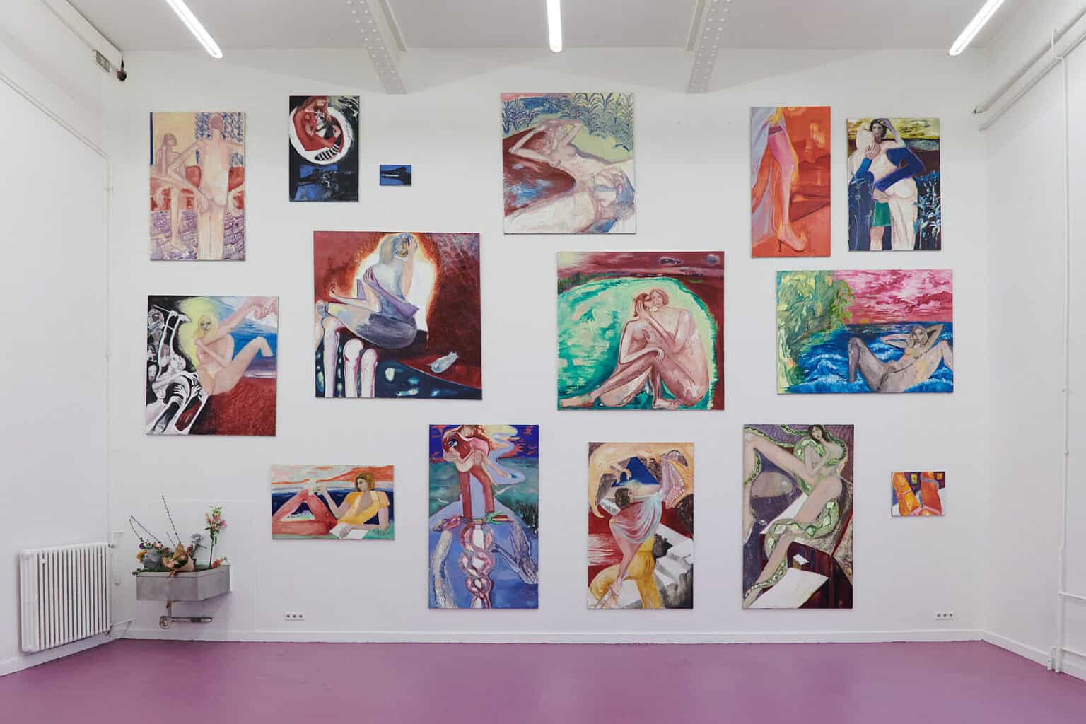 Foto: Ansicht eines Raumes mit weißen Wänden und rosa-lilanem Boden. An der Wand befindet sich ein Waschbecken, in dem sich Blumen befinden. Die gesamte Wand ist, bis unter die Decke, mit Leinwandarbeiten im Klein-, Mittel- und Großformat behangen.