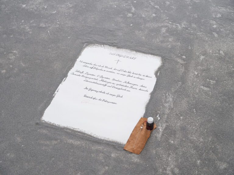 Foto: Detailaufnahme eines Bogens Papier mit einem handschriftlichen Text. Das Papier liegt auf einer Fläche mit grauem Sand. Neben dem Papier liegt ein braunes Stück Leder mit einem kleinen Fass und einem Glasfüller.