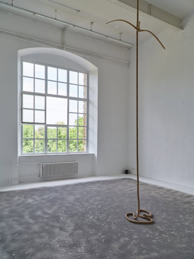 Foto: Aufnahme eines Ausstellungsraums mit weißen Wänden und weißem Boden und Rundbogenfenster. Der Boden ist mit grauem Sand aufgestreut. In der Mitte des Raums hängt ein Seil von der Decke bis zum Boden. An dem Seil hängt ein Holzbogen.