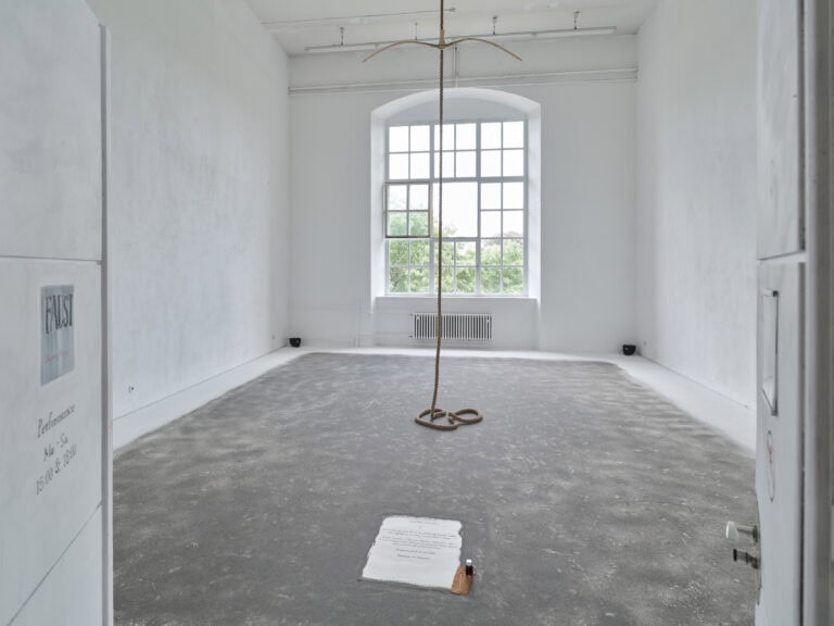 Foto: Aufnahme eines Ausstellungsraums mit weißen Wänden und weißem Boden und Rundbogenfenster. Der Boden ist mit grauem Sand aufgestreut. In der Mitte des Raums hängt ein Seil von der Decke bis zum Boden. An dem Seil hängt ein Holzbogen. Auf dem Boden liegt ein Bogen Papier mit einem handschriftlichen Text.