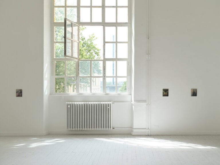 Foto: Aufnahme eines Ausstellungsraums mit weißen Wänden und weißen Dielenboden und Fenster. An der Rückwand sind drei kleinformatige Gemälde angebracht.