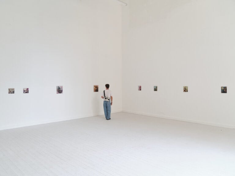 Foto: Aufnahme eines Ausstellungsraums mit weißen Wänden und weißen Dielenboden und Fenster. An der Rückwand hängen acht kleinformatige Gemälde. Eine Person steht in der Raumecke und betrachtet die Bilder.