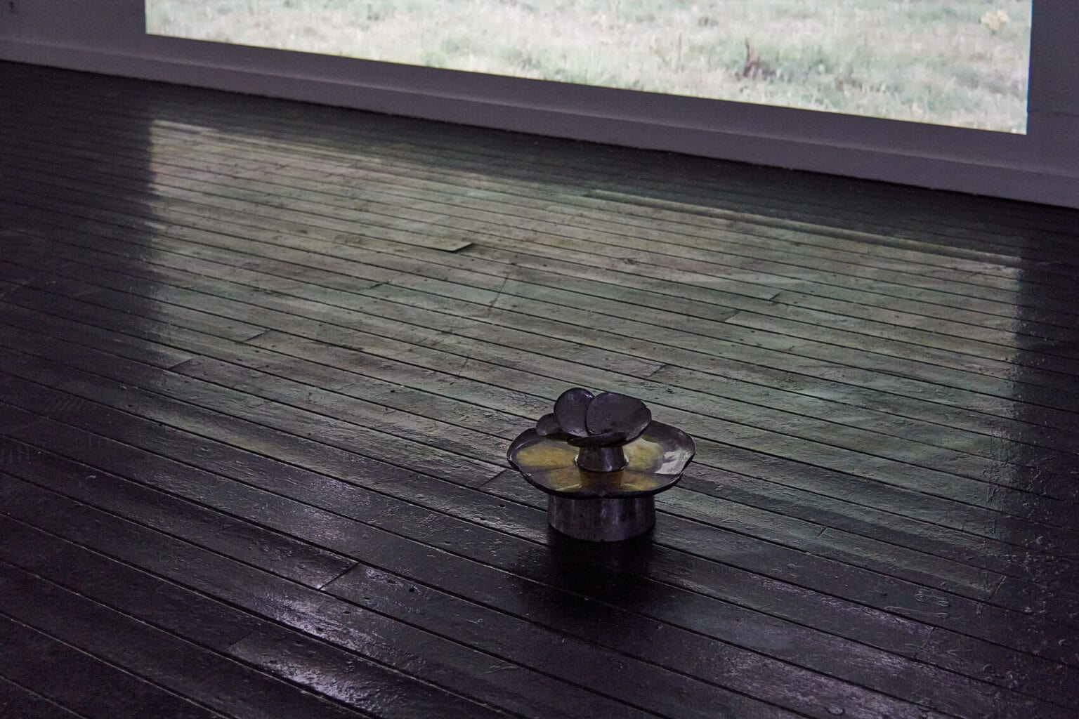 Foto: Ein abgedunkelter Raum mit Dielenboden. Auf dem Boden steht ein Objekt. Der Boden und das Objekt reflektiert weiß-grünliches Licht, das an die Wand geworfen wird.