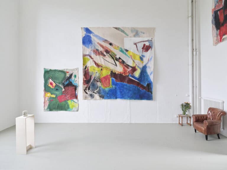 Foto: Aufnahme eines Ausstellungsraums mit weißen Wänden und grauem Boden. An der Wand hängen drei großformatige Stoffe mit abstrakter bunter Malerei. Am linken Rand steht ein weißes u-förmiges Tischelement mit einem aufstellten Buch. Am rechten Rand steht eine braune Ledercouch und ein Beistelltisch mit einem Strauss Blumen.