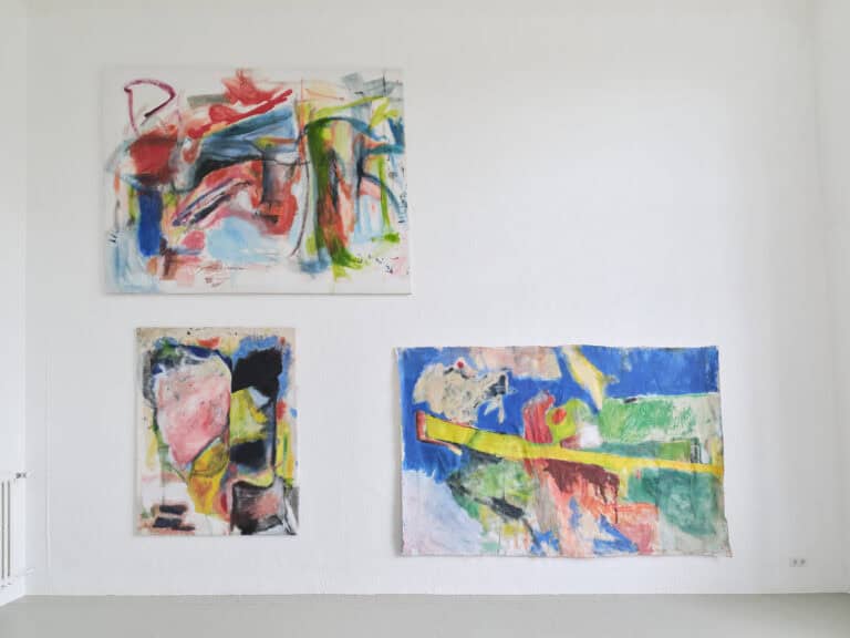 Foto: Aufnahme eines Ausstellungsraums mit weißen Wänden und grauem Boden. An der Wand hängen drei großformatige Stoffe mit abstrakter bunter Malerei.