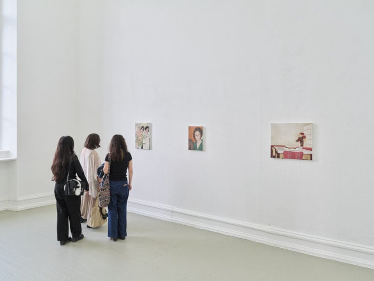 Foto: Aufnahme eines Ausstellungsraums mit weißen Wänden und grauem Boden. An der Wand hängen drei Gemälde. Eine Gruppe von drei Personen betrachtet die Bilder.