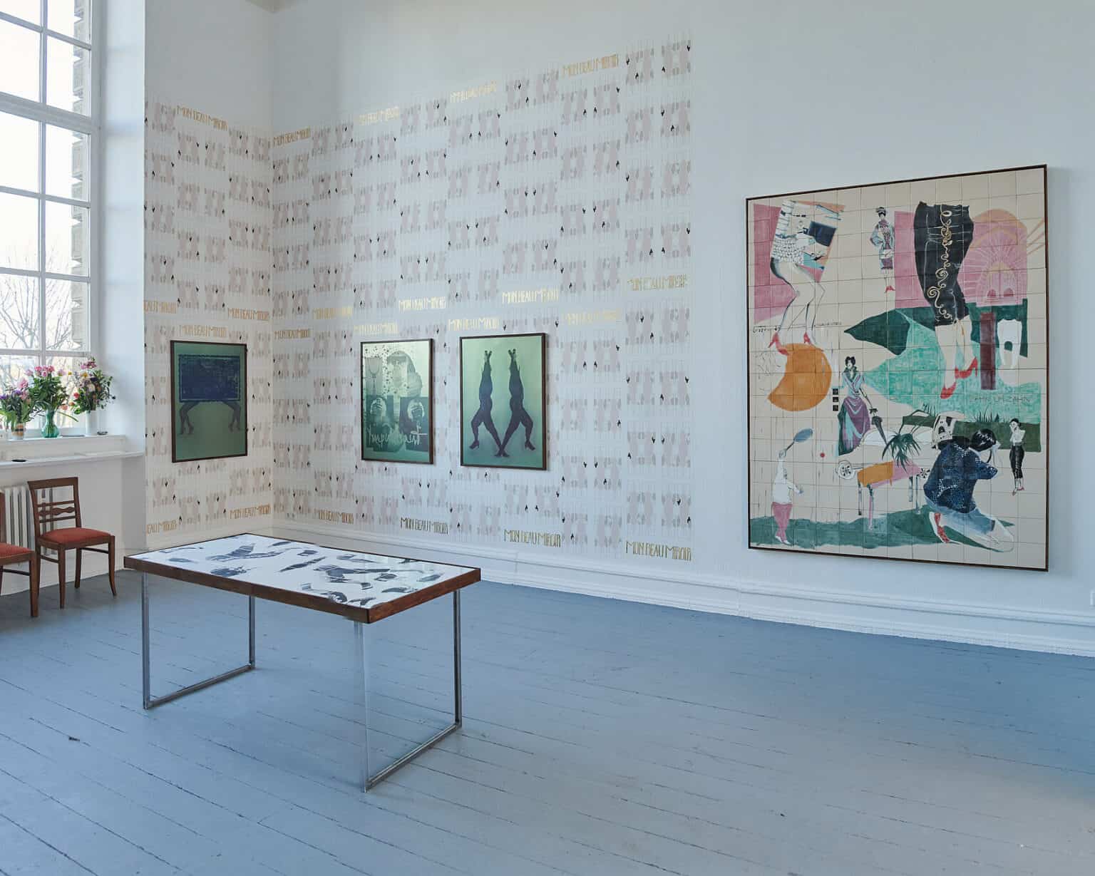 Foto: Ansicht eines Raumes mit weißen Wänden, grauem Dielenboden und einem großen Fenster. Ein Teil der Wand ist mit einer gemusterten Tapete oder Bemalung versehen. Auf der Fensterbank stehen Blumensträuße. An der Wand hängen insgesamt vier mittel- und großformatige Objekte. Im Raum steht ein Tisch, vor der Fensterbank zwei Stühle.