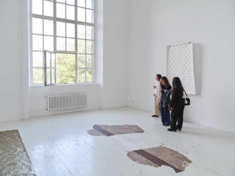 Foto: Aufnahme eines Ausstellungsraums mit weißen Wänden und weißen Dielenboden und Fenster. An der Rückwand hängen eine Papierarbeit. Auf dem Boden sind weitere Malereien zu sehen. Eine Gruppe von drei Personen betrachtet die Werke.