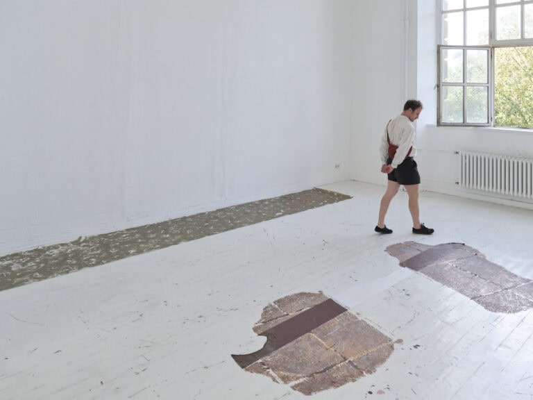Foto: Aufnahme eines Ausstellungsraums mit weißen Wänden und weißen Dielenboden und Fenster. Auf dem Boden sind Malereien zu sehen. Eine Person geht durch den Raum und betrachtet die Werke.