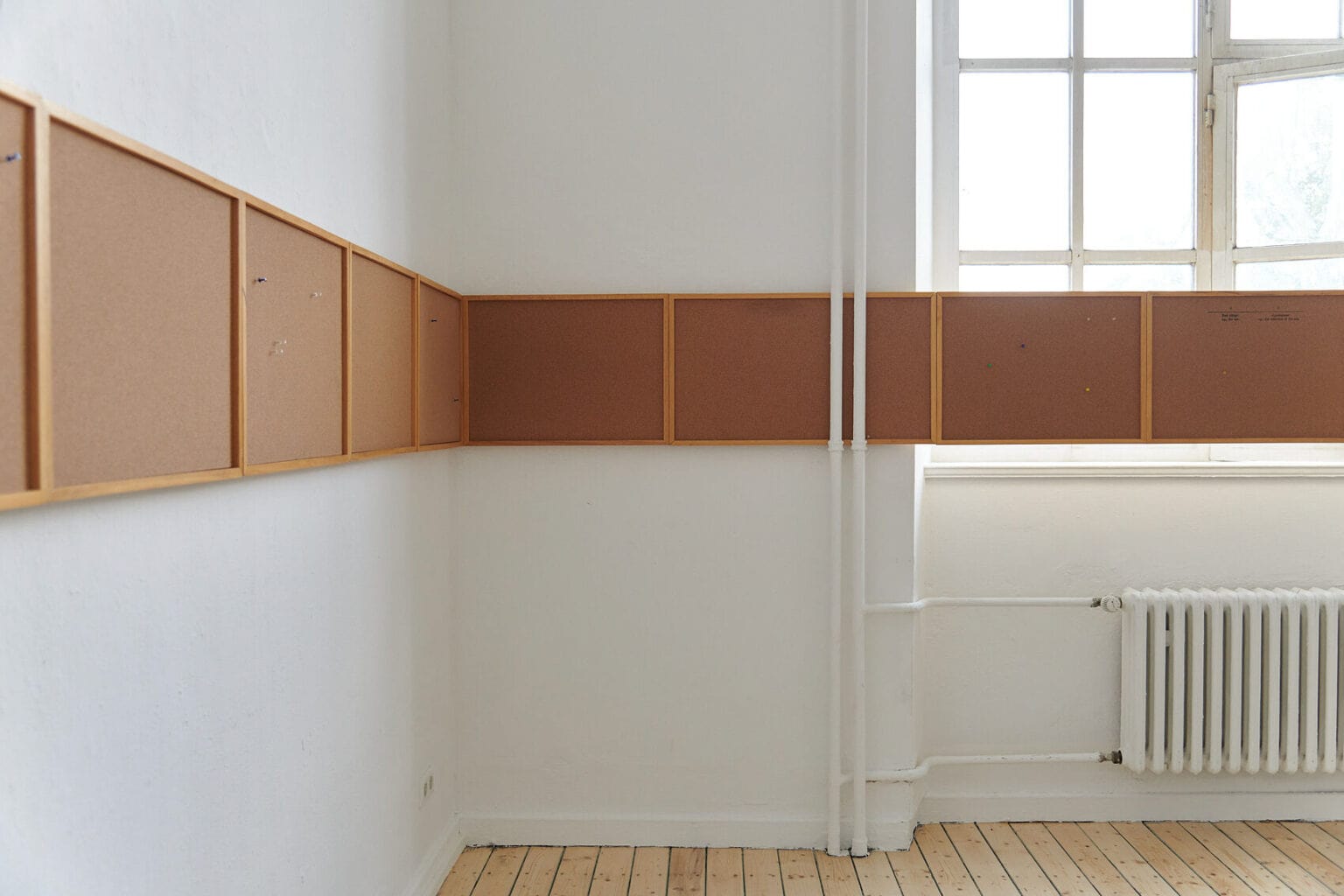 Foto: Ein Raum mit weißen Wänden, Dielenboden und einem großen Fenster. Die Wände des Raumes sind von einer durchgehenden Reihe aus flachen Tafeln bedeckt, die sich auch über das Fenster erstreckt.