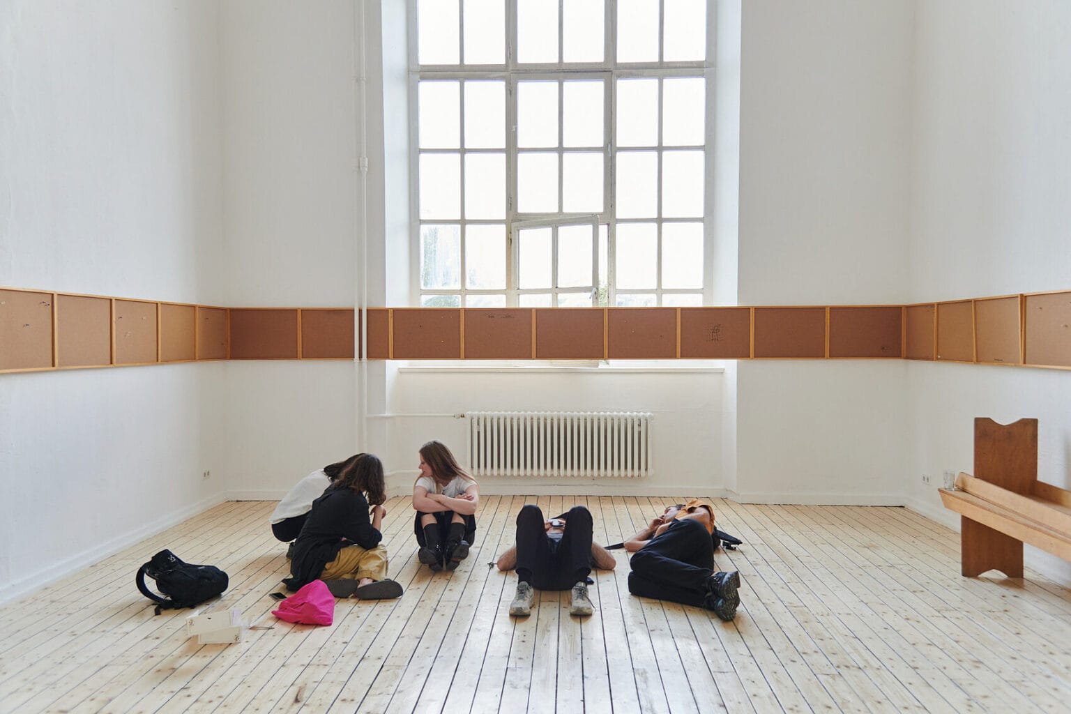 Foto: Ein Raum mit weißen Wänden, Dielenboden und einem großen Fenster. Auf dem Biden des Raume liegen und sitzen Menschen. Die Wände des Raumes sind von einer durchgehenden Reihe aus flachen Tafeln bedeckt, die sich auch über das Fenster erstreckt.