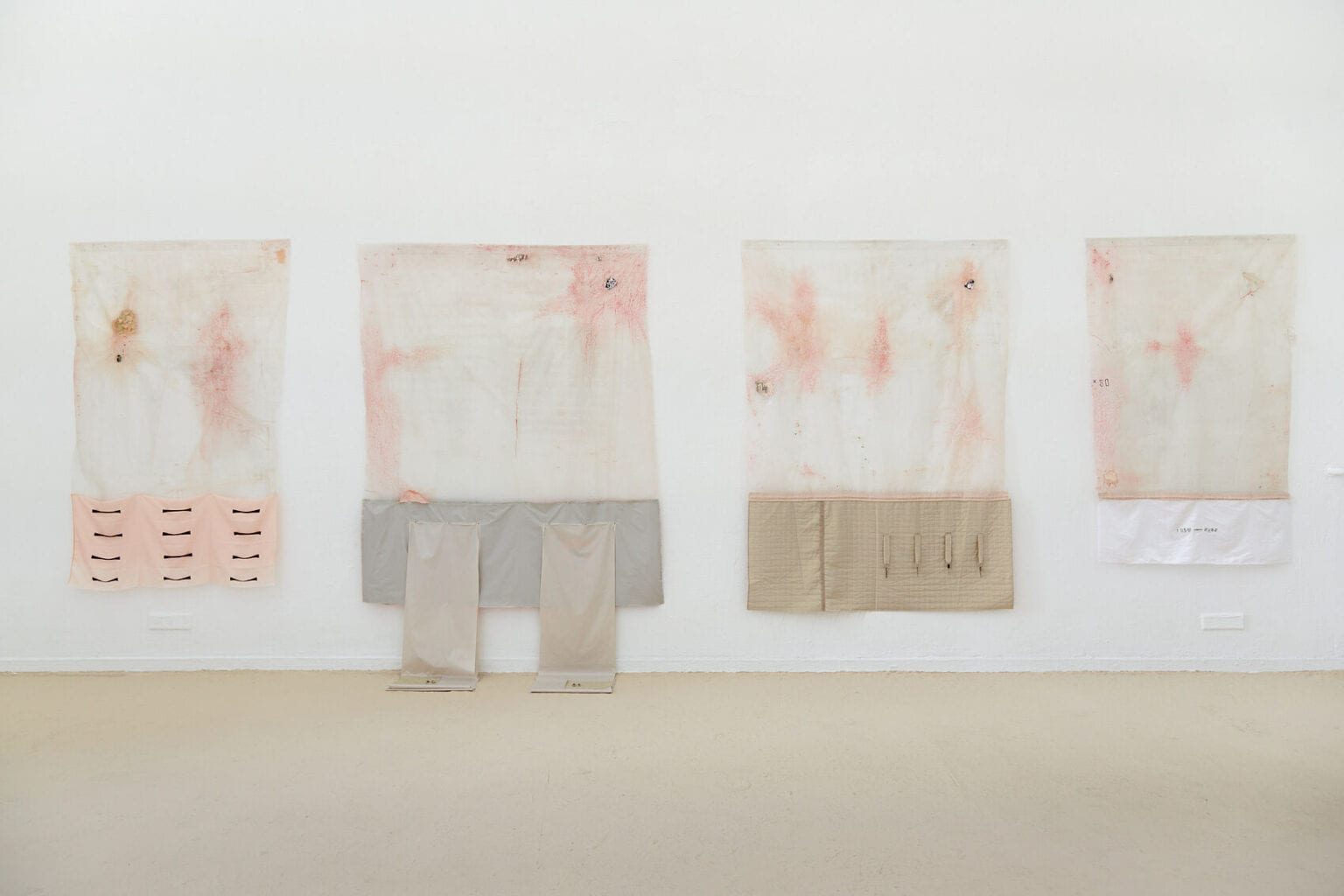 Foto: Ein Raum mit weißen Wänden und beigem Boden. An der Wand befinden sich vier große, quadratische Objekte in unterschiedlicher Größe, die auf gleicher Oberkante hängen. Teile eines Objektes liegen auf dem Boden auf.