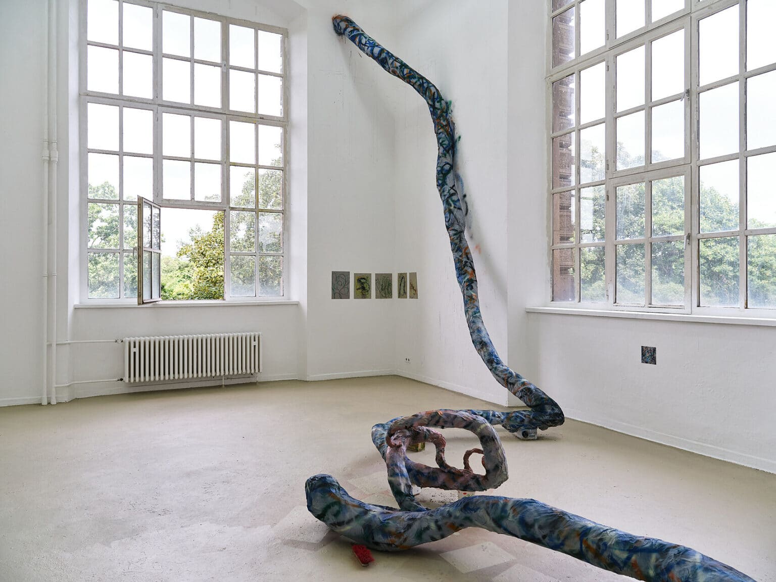 Foto: Ansicht eines Raumes mit weißen Wänden, beigem Boden und zwei Fenstern. An den Wänden des Raumes befinden sich kleinformatige flache Objekte. Über den Boden und eine Wand des Raumes erstreckt sich ein längliches, buntes Objekt.