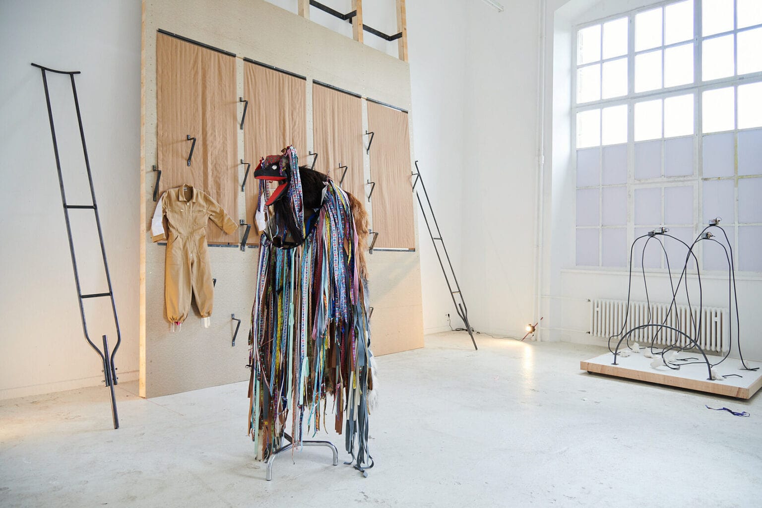 Foto: Ein Raum mit weißen Wänden, zum Teil abgedunkelten Fenstern und hellem Boden. Im Raum verteilt befinden sich mehrere Objekte und eine große Stellwand, die an der Wand lehnt. Auf der Stellwand sind Haken, Textilien und ein Overall montiert. Links und rechts von der Stellwand befinden sich zwei längliche Konstruktionen.