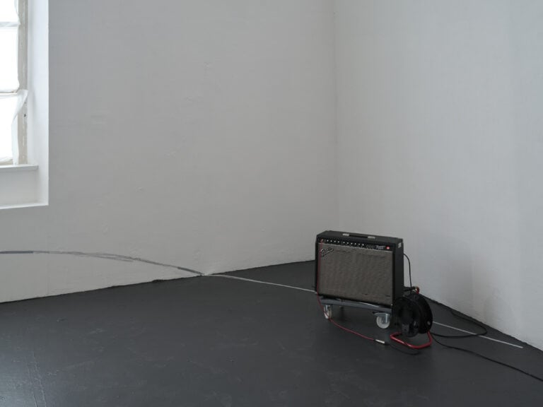 Foto: Aufnahme einer Raumecke in einem Ausstellungsraum mit weißen Wänden, dunklem Boden und angeschnittenem Fenster. In der Ecke steht eine Kunstinstallation bestehend aus einem Musikverstärker auf einem Rollbrett und eine Kabeltrommel.