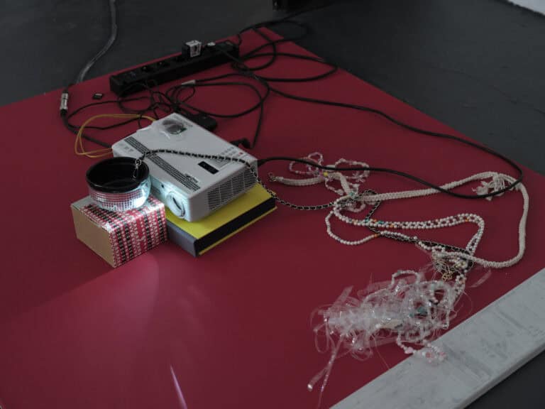 Foto: Detailaufnahme einer Kunstinstallation. Auf einer roten Fläche liegen diverse Gegenstände, z.B. ein Beamet, eine Mehrfachsteckdose, Ketten, Folienstreifen.