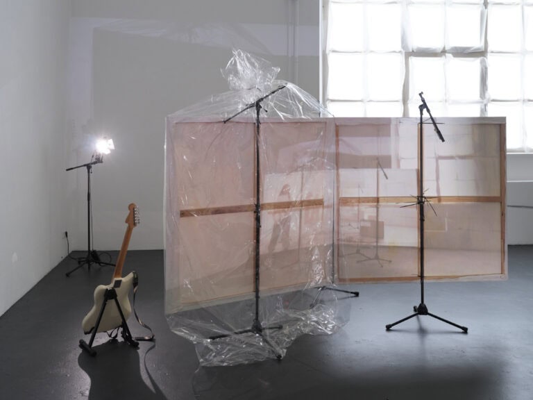 Foto: Aufnahme eines Ausstellungsraums mit weißen Wänden, dunklem Boden und großem Fenster. Im Raum ist eine mehrteilige Rauminstallation aufgebaut, bestehend aus Leinwänden, die als Projektionsfläche dienen, Mikrophonen, einer E-Gitarre auf einem Ständer. Ein Teil der Installation ist in Klarsichtfolie verpackt.