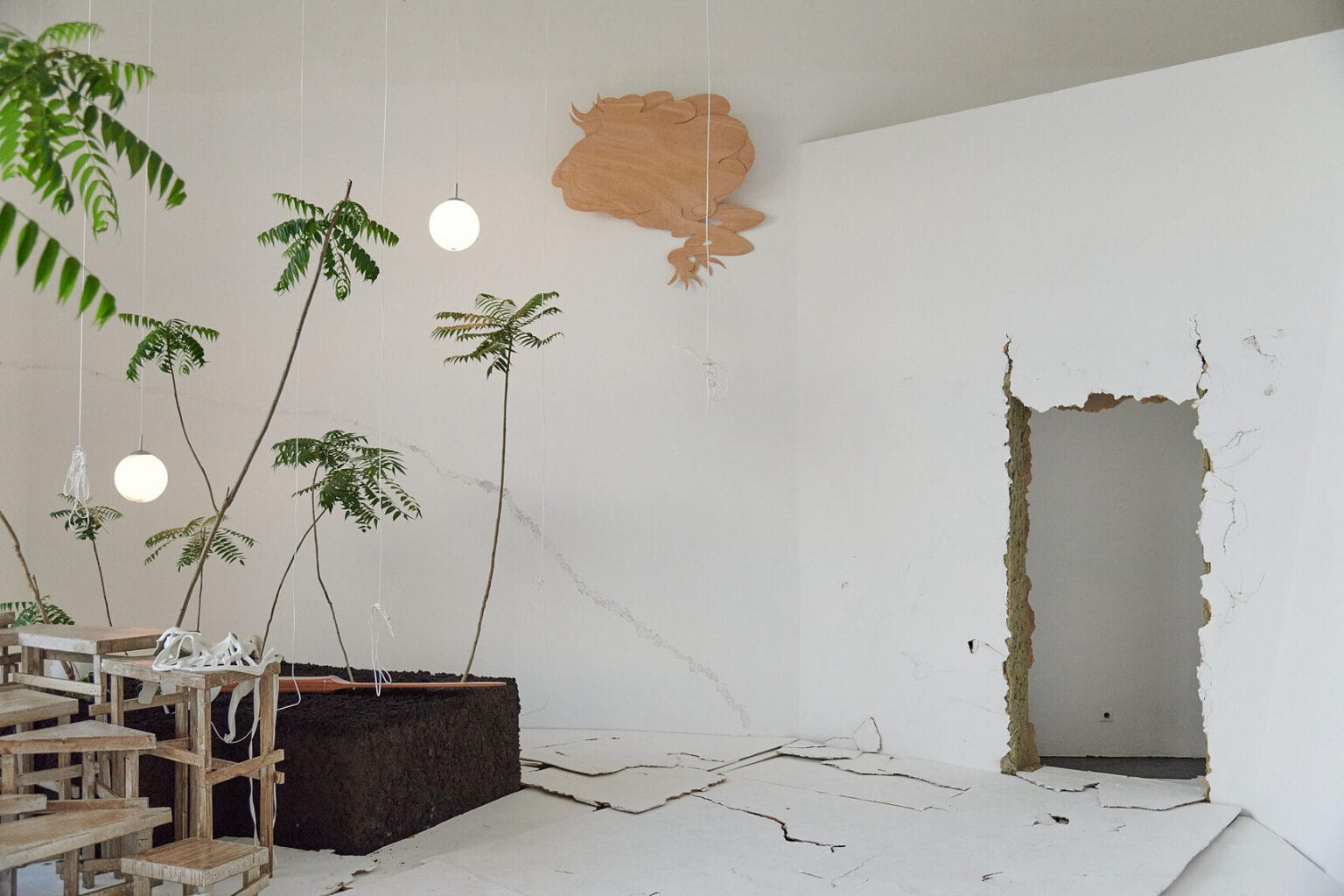 Foto: Ein Raum mit weißen Wänden und rahmenlosem Durchbruch in der Größe einer Tür. Die Wand um die Türöffnung ist rissig. Im Raum befindet sich eine Konstruktion auf verschiedenen Materialien, darunter Schüre, Lampen und Pflanzen. Oben an der Wand hängt ein Objekt und an anderer Stelle wird ein Bild an eine Wand projiziert. Der Boden des Raumes ist mit verschieden großen, hellen Platten bedeckt, die zum Teil übereinander liegen und eine unebene, rissige Oberfläche erzeugen.