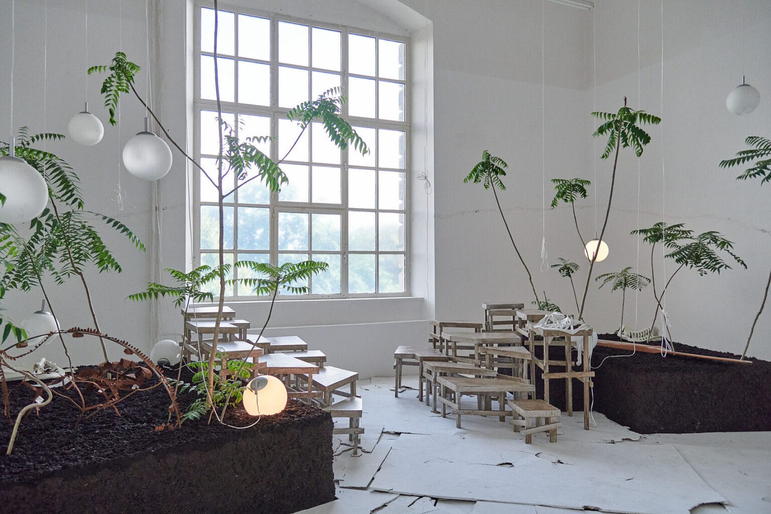 Foto: Ein Raum mit weißen Wänden und großen Fenster. Im Raum befindet sich eine große Konstruktion auf verschiedenen Materialien, darunter verschieden große Podeste, die Stufenartig angeordnet sind, ein großer mit Erde gefüllter Kübel in dem sich Blumen befinden und von der Decke herabhängende Lampen und Schnüre. Der Boden des Raumes ist mit verschieden großen, hellen Platten bedeckt, die zum Teil übereinander liegen und eine unebene Oberfläche erzeugen.