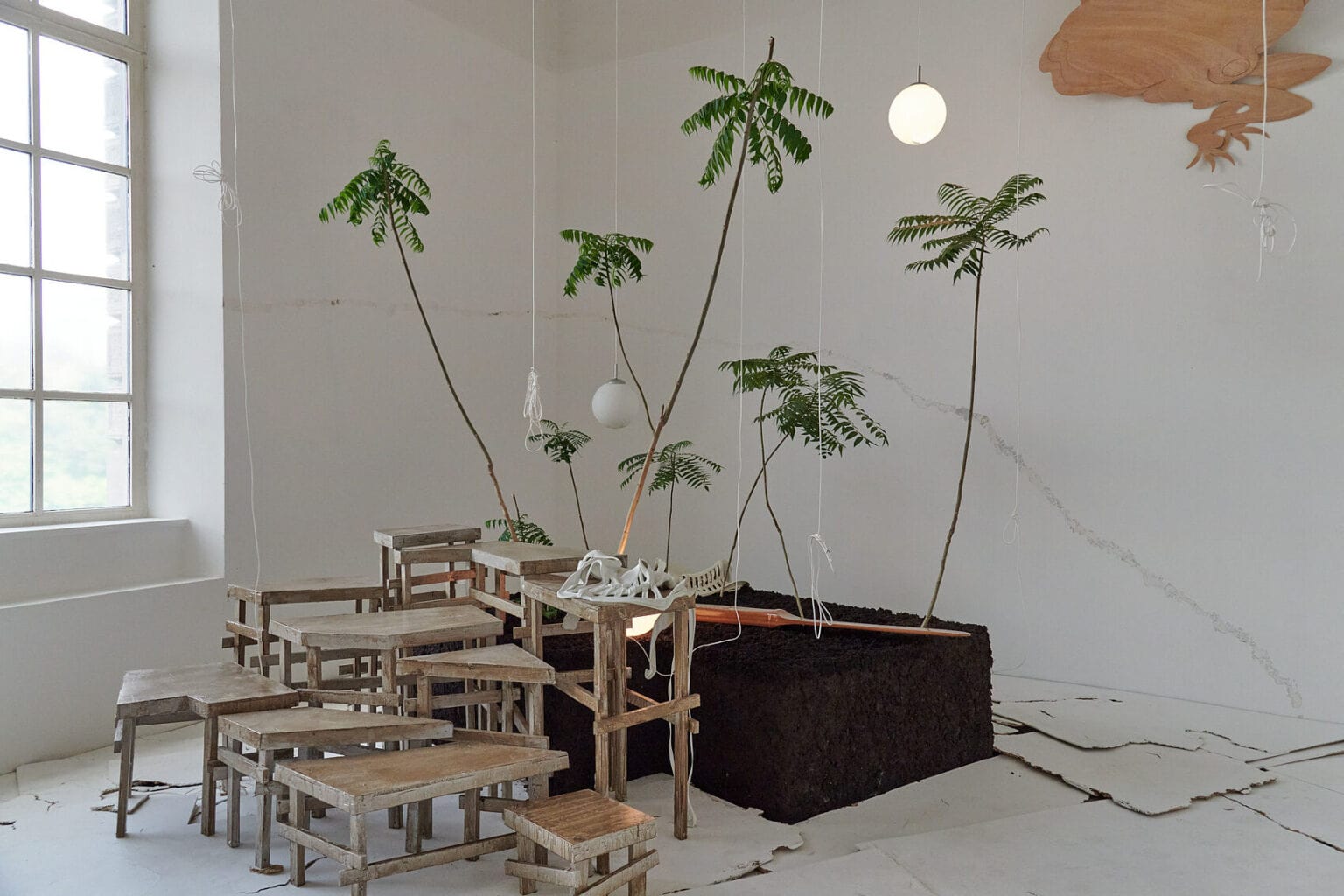 Foto: Ein Raum mit weißen Wänden und Fenster. Im Raum befindet sich eine große Konstruktion auf verschiedenen Materialien, darunter verschieden große Podeste, die Stufenartig angeordnet sind, ein großer mit Erde gefüllter Kübel in dem sich Blumen befinden und von der Decke herabhängende Lampen und Schnüre. Der Boden des Raumes ist mit verschieden großen, hellen Platten bedeckt, die zum Teil übereinander liegen und eine unebene Oberfläche erzeugen.