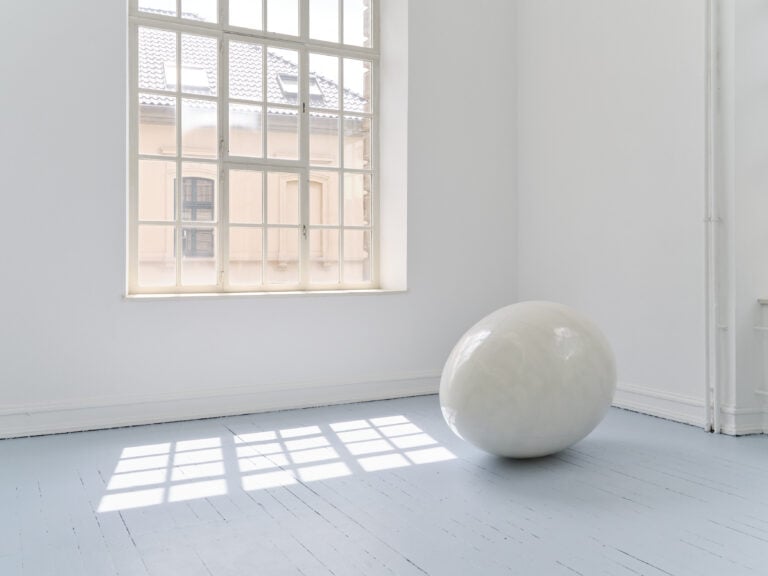 Foto: Aufnahme eines Ausstellungsraums mit weißen Wänden und grauem Boden und Fenster. Im Raum liegt eine großformatige weiß glänzende Ei-Skultpur