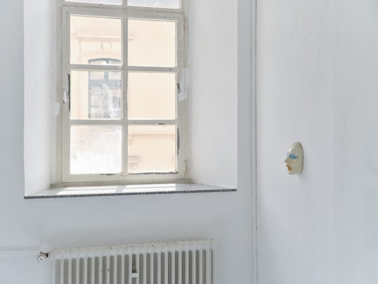 Foto: Aufnahme eines Ausstellungsraums mit weißen Wänden und Fenster. An der rechten Wand ist eine weiße Maske angebracht.