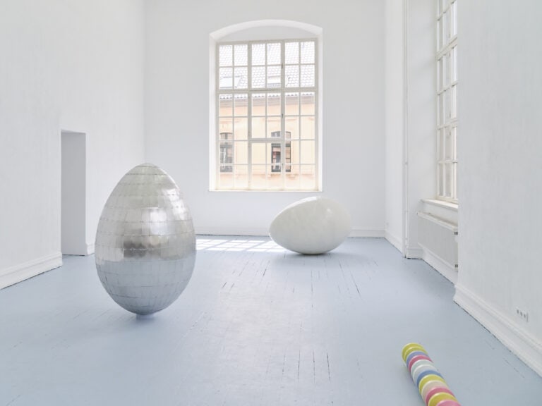 Foto: Aufnahme eines Ausstellungsraums mit weißen Wänden und grauem Boden und Rundbogenfenster. Im Raum stehen zwei großformatige Ei-Skultpuren, eine aus silbernen Platten zusammengesetzt, eine weiß glänzend. Im vorderen Bereich liegt eine Reihe bunter Perlen.