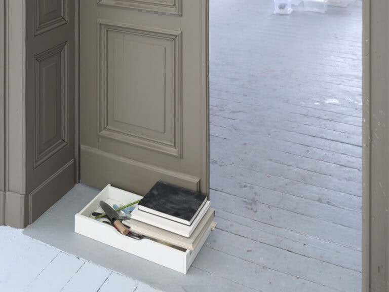 Foto: Detailaufnahme einer Kunstinstallation. Eine Schublade mit diversen Gegenständen gefüllt steht vor einem Türdurchgangs auf grauem Dielenboden.