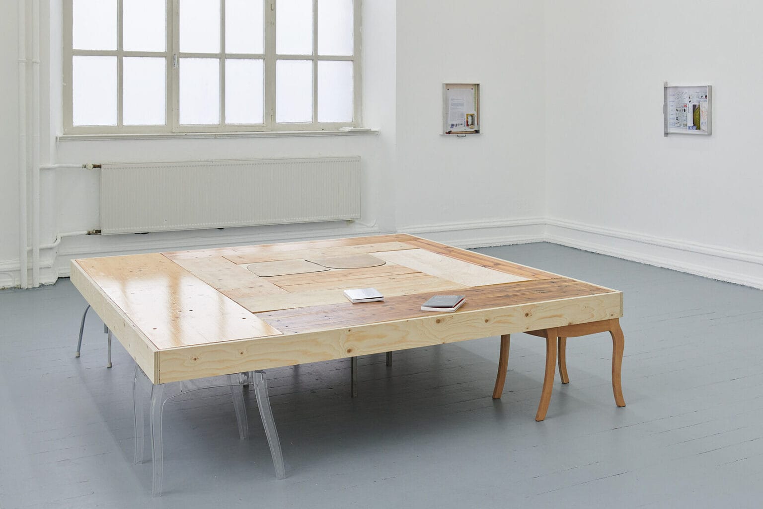 Foto: Ansicht eines Raumes mit weißen Wänden, grauem Dielenboden und einem großen Fenster. An den Wänden befinden sich zwei Objekte. Im Raum steht ein niedriger, aus verschiedenfarbigen Holzleisten und Platten bestehender, Tisch. Der Tisch wird an seinen vier Ecken von hockeraktigen, jeweils vierbeinigen Konstruktionen getragen, die aus unterschiedlichen Materialien gefertigt sind. Auf dem Tisch liegen zwei Hefte.