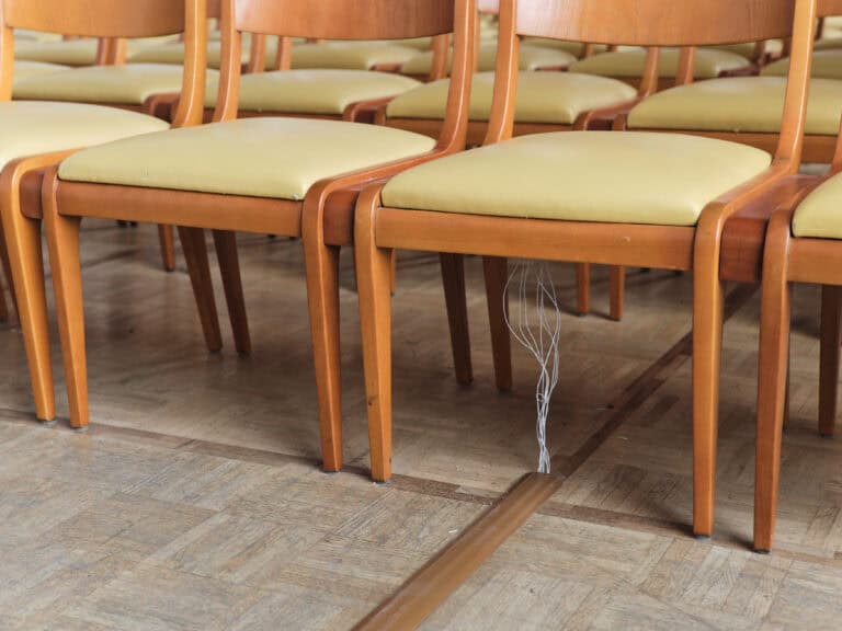 Foto: Detailaufnahme einer Reihe Holzstühle mit beigen Sitzflächen. In einem Kabelkanal laufen dünne weiße Kabel zu der Unterseite eines Stuhls.