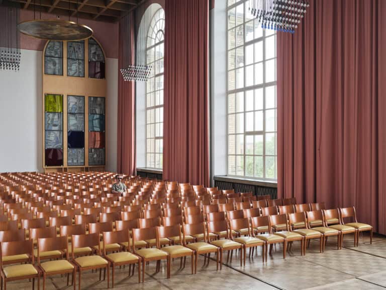Foto: Aufnahme einer Halle mit großen Rundbogenfenstern und langen Vorhängen. In der Halle stehen viele Reihen mit Holzstühlen und beigen Sitzflächen. Mittig sitzt eine Person auf einem der Stühle.