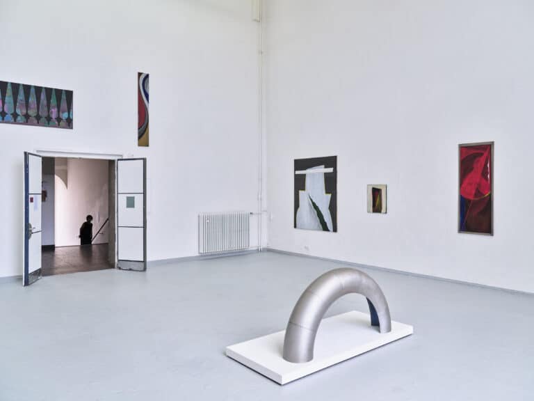 Foto: Aufnahme eines Ausstellungsraums mit weißen Wänden, grauem Boden und einer geöffneten Tür mit zwei Flügeln. An den Wänden hängen abstrakte Gemälde. Auf dem Boden steht ein silbernes Bogenelement auf einem weißen Podest.