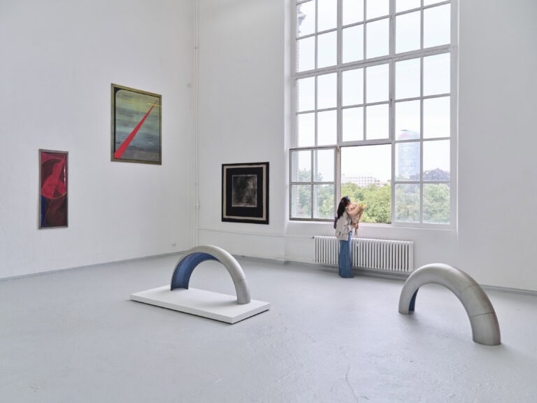 Foto: Aufnahme eines Ausstellungsraums mit weißen Wänden, grauem Boden und großem Fenster. An den Wänden hängen abstrakte Gemälde. Auf dem Boden stehen zwei silberne Bogenelemente, einer auf einem weißen Podest. An dem Fenster steht eine Person mit einem Blumenstrauß.