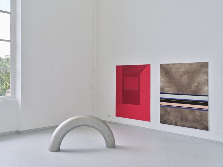 Foto: Aufnahme eines Ausstellungsraums mit weißen Wänden, grauen Boden und angeschnittenem Fenster. An den Wänden hängen zwei großformatige abstrakte Gemälde. Auf dem Boden steht ein silbernes Bogenelement.