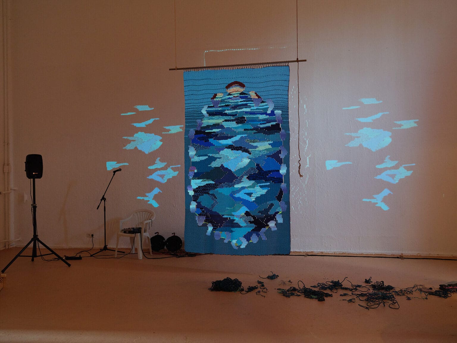 Foto: Ansicht eines abgedunkelten Raumes mit weißen Wänden und farbigem Boden. Auf dem Boden liegt eine Gruppe von Objekten. Links im Raum steht Technik, darunter ein Lautsprecher und ein Mikrofonständer, Kabel und ein Stuhl. Von der Decke hängt ein an einer Leiste befestigtes Textil, dass von einer Projektion angestrahlt wird, die auch die umliegende Wand anleuchtet.