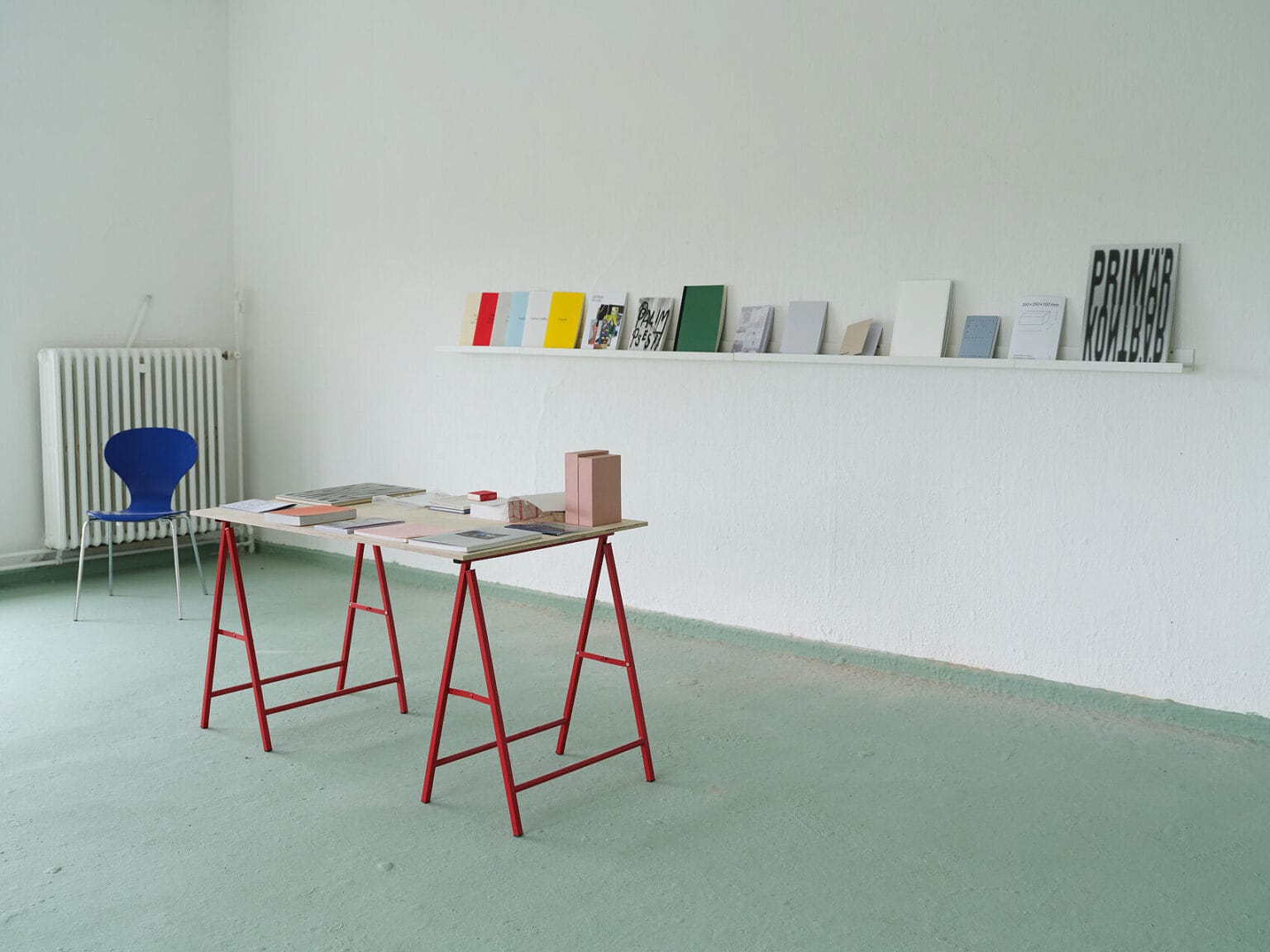 Foto: Ansicht eines Raume mit weißen Wänden und grünem Boden. An der Wand befindet sich eine Leiste, auf der sich eine Reihe unterschiedlicher Bücher befinden. Im Raum steht ein Stuhl und ein Tisch. Auf dem Tisch befinden sich ebenfalls Bücher und Objekte.