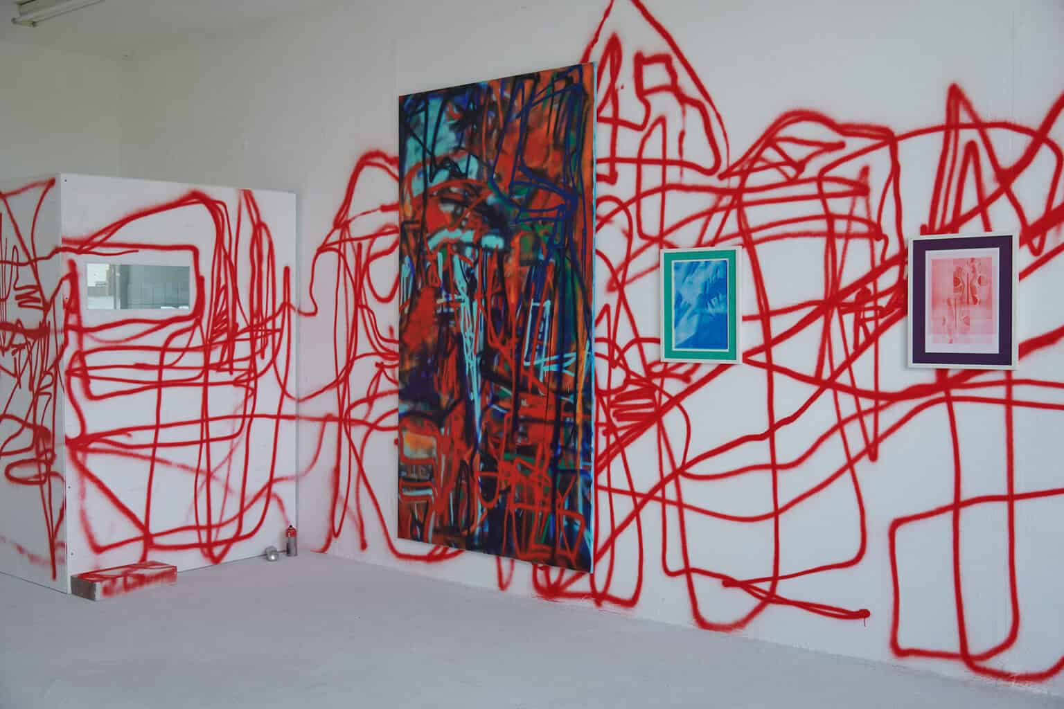 Foto: Ein Raum mit weißen Wänden und grauem Boden. Die Wände sind mit einem Muster aus roten Linien besprüht. Auf einer dieser Wände hängen Arbeiten im Groß- und Kleinformat.