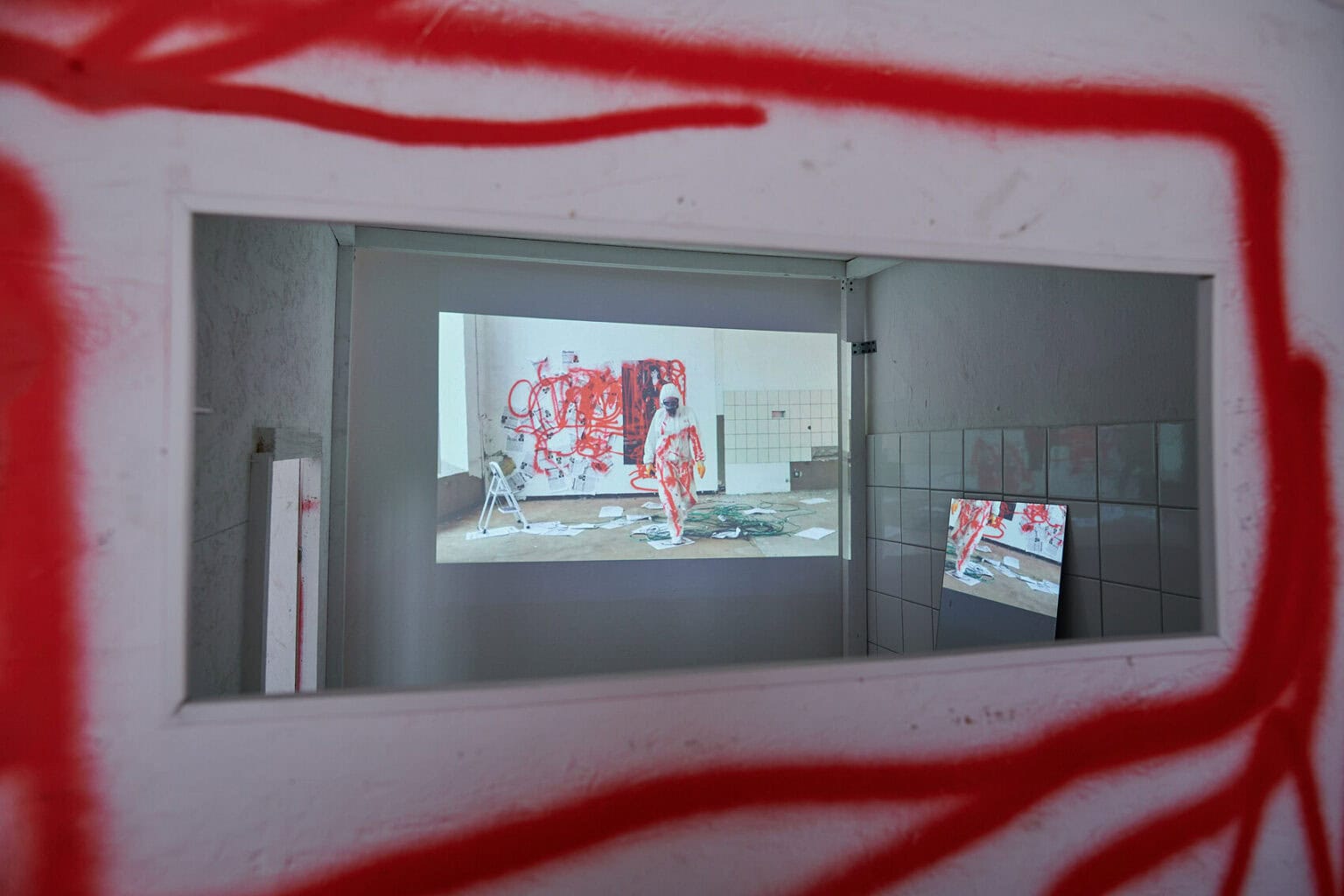 Foto: Ein kleiner, halbdunkler Raum mit weißen Wänden und Fliesen. Im Raum befinden sich eine große und eine kleine Projektion, die ein ähnliches Motiv zeigen.